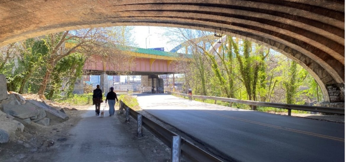 Photo looking under and through an overpass. People are using a trail that is separated from the roadway. Sun is shining. Location is Falls Road at North Avenue (Jones Falls Trail).